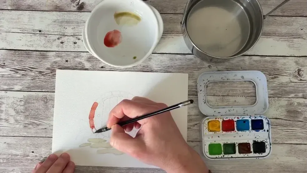 Étape d'un tutoriel vue de dessus, une main peint à l'aquarelle sur une feuille, à côté d'une palette de godets et d'un pot d'eau.